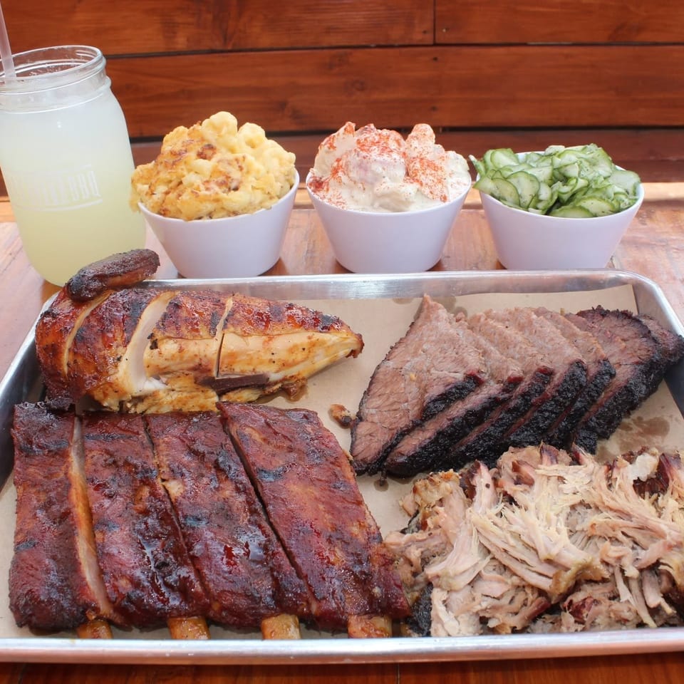 Barbecue platter with ribs, brisket, pulled pork, roasted chicken, side dishes, and lemonade on a wooden table.
