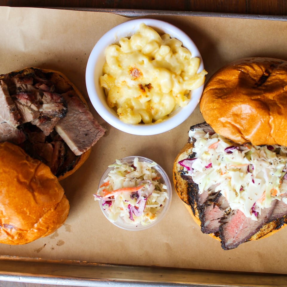 Overhead view of barbecue sandwiches with sliced brisket, macaroni and cheese, and coleslaw on a paper-lined tray.