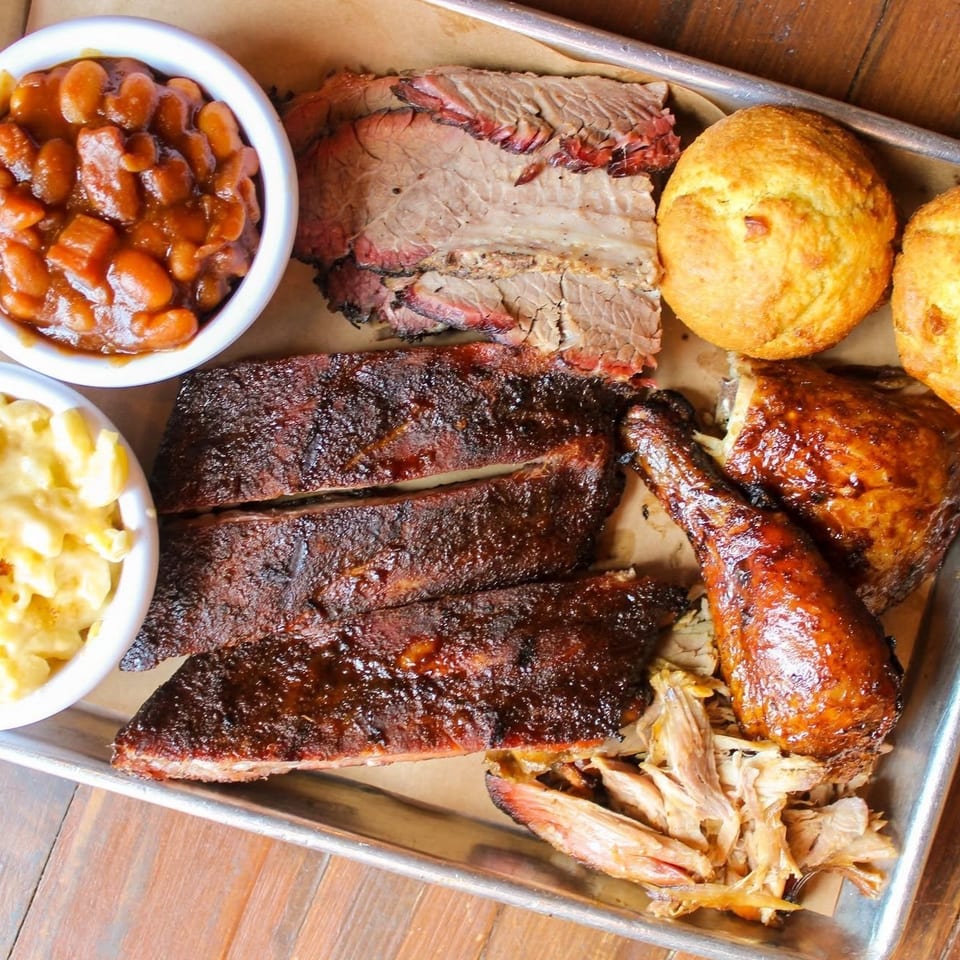 Barbecue platter with sliced brisket, ribs, glazed chicken, pulled pork, baked beans, macaroni and cheese, and cornbread muffins.