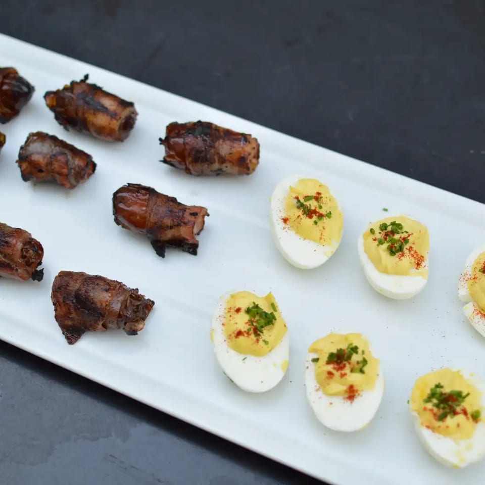 White platter of grilled bacon-wrapped bites and deviled egg halves garnished with herbs and paprika.