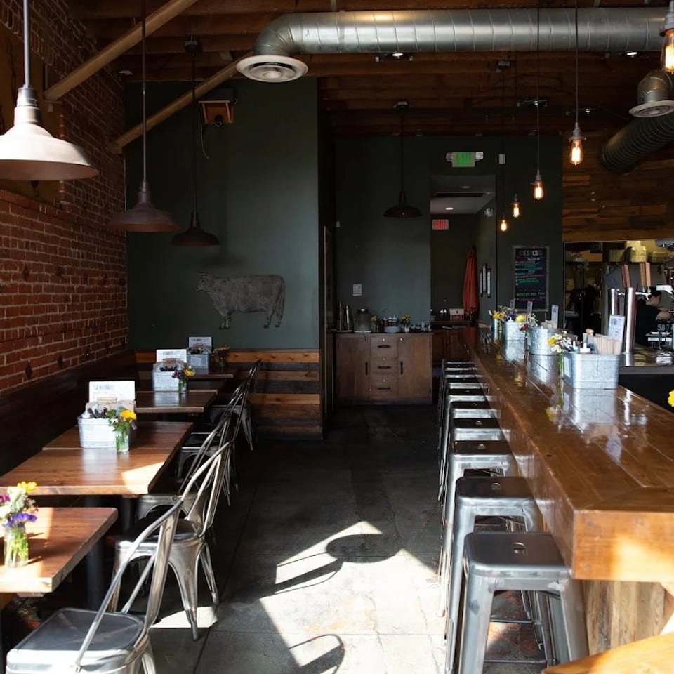 Industrial-style restaurant interior with exposed brick walls, wooden tables, metal stools, hanging lights, and a long polished bar.