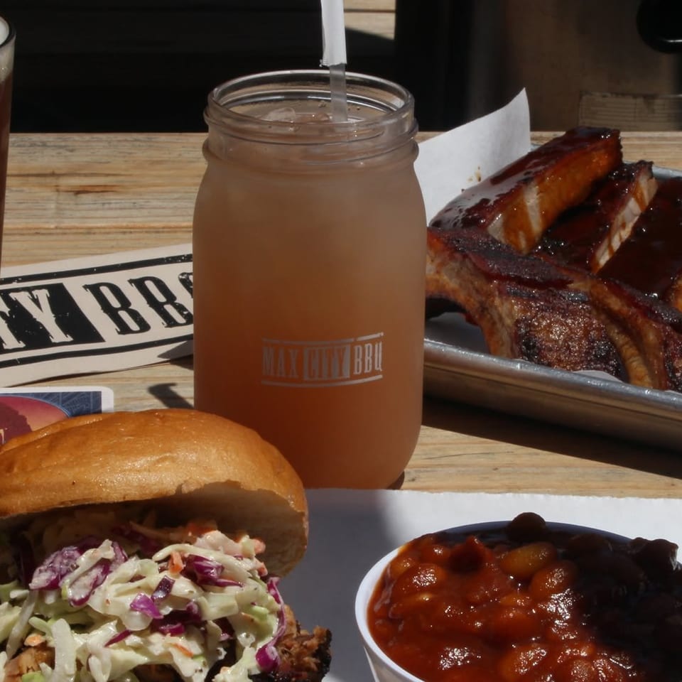 Barbecue spread with sauced ribs, pulled pork sandwich topped with coleslaw, baked beans, beer, and a mason-jar drink.