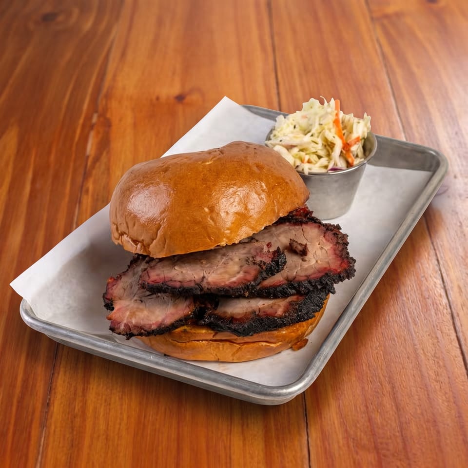 Smoked meat sandwich on a glossy bun with barbecue bark, served alongside creamy coleslaw on a metal tray.