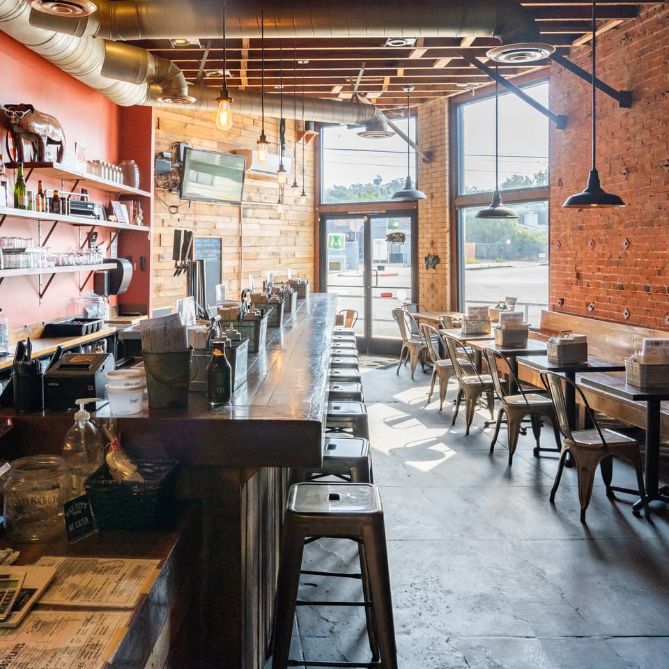 Industrial-style restaurant interior with exposed brick, wood walls, bar seating, tables, metal chairs, and large sunlit windows.