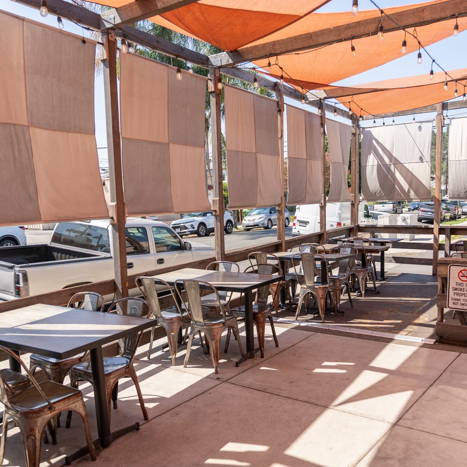 Outdoor restaurant patio with metal tables, chairs, orange shade canopies, string lights, and fabric privacy screens.