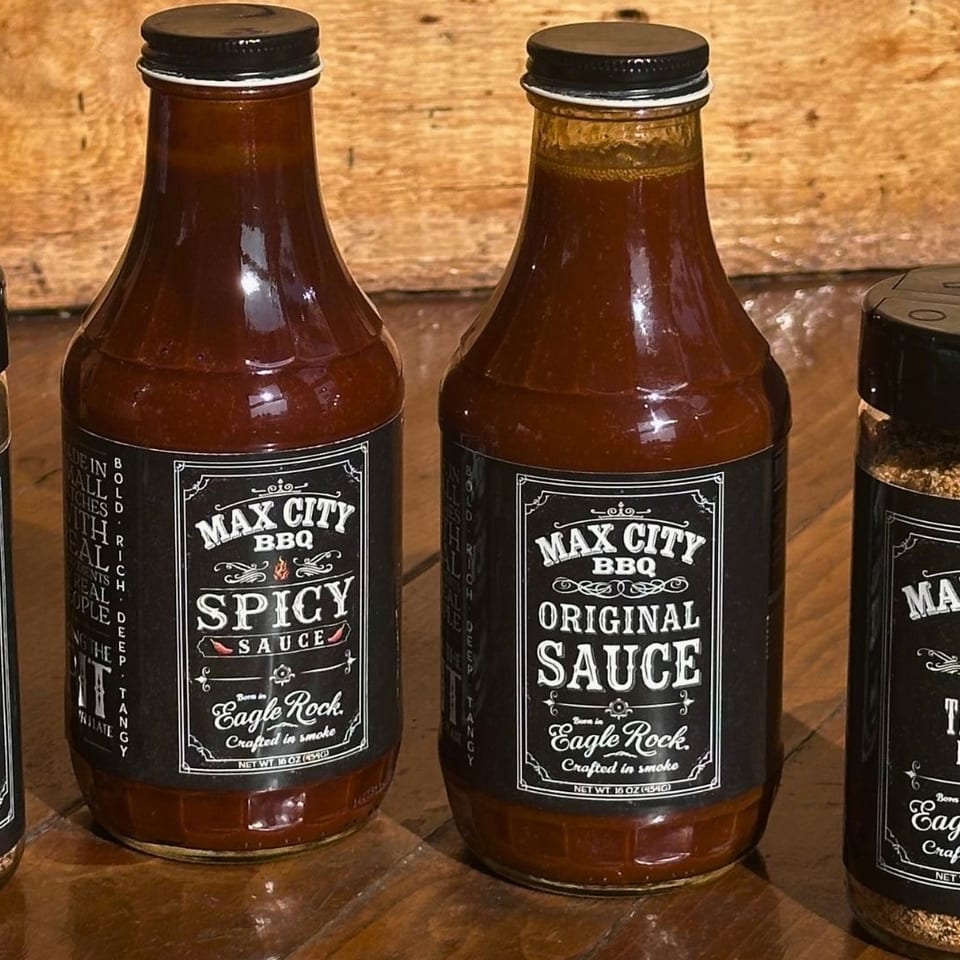 Four Max City BBQ products—two sauce bottles and two spice rub shakers—stand on a wooden table against a rustic wood backdrop.