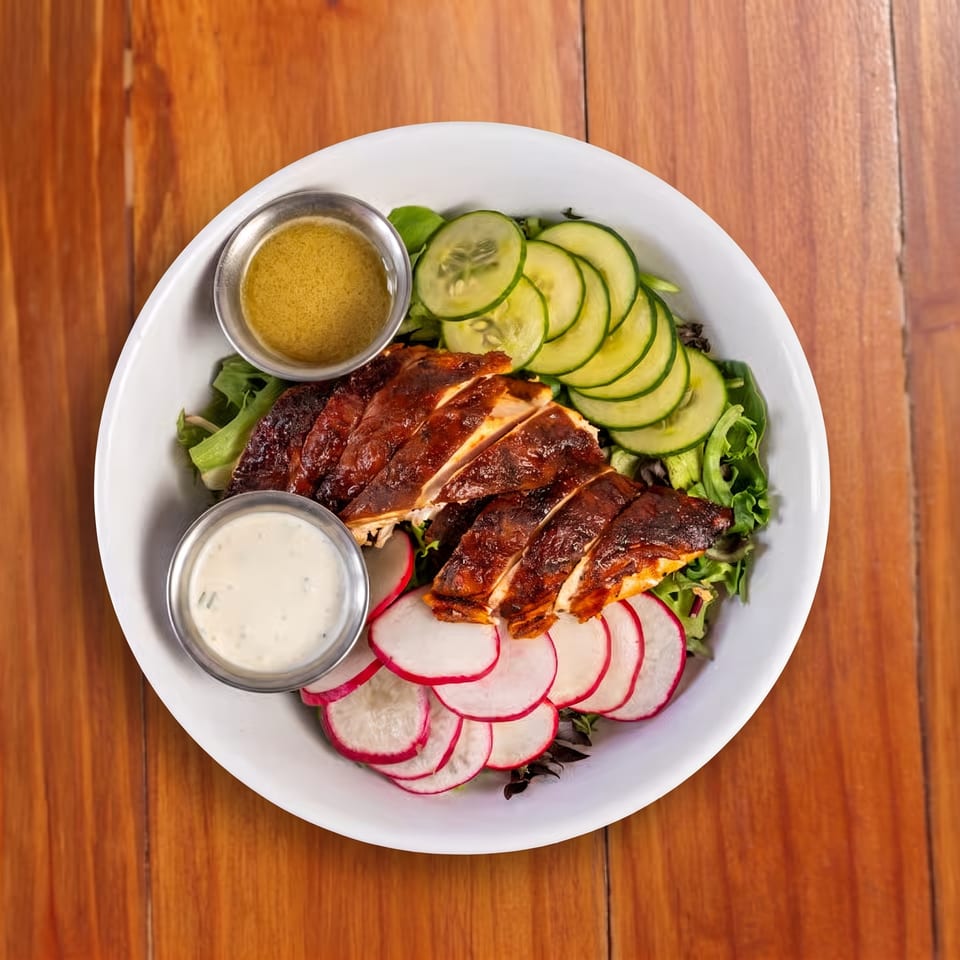 Grilled chicken salad with cucumbers, radishes, greens, and two dipping sauces on a white plate atop a wooden table.