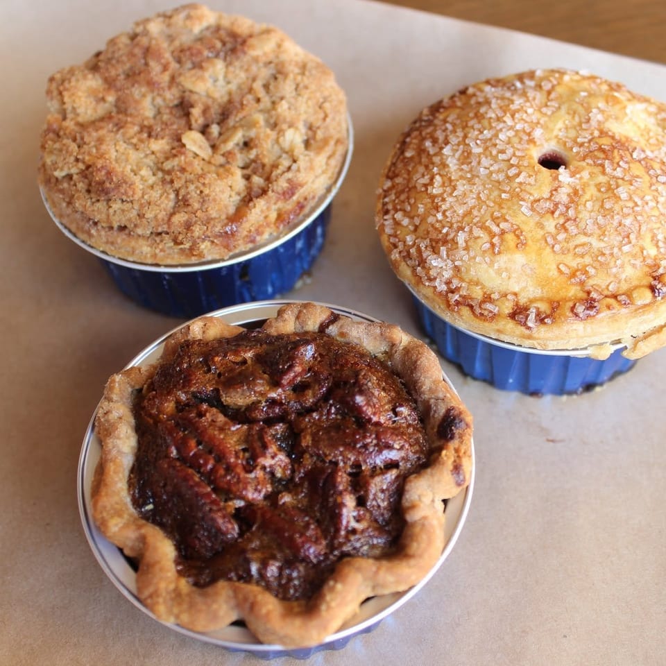 Three rustic fruit pastries in blue baking tins, including crumb-topped, sugar-glazed, and dark caramelized varieties.