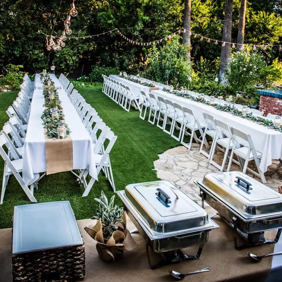 Elegant outdoor wedding reception with white tables, folding chairs, greenery, string lights, and covered buffet chafing dishes.