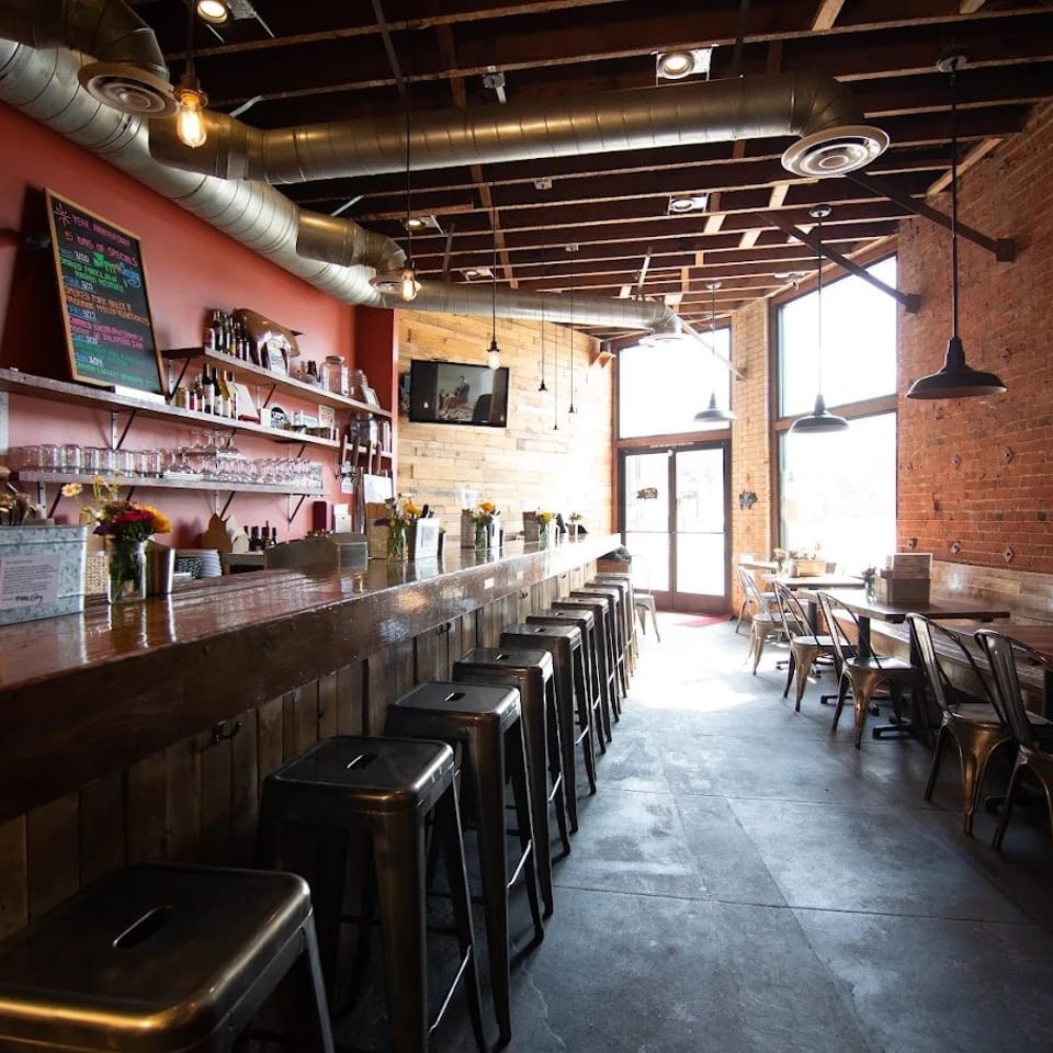 Industrial-style barbecue restaurant interior with a long wooden bar, metal stools, exposed brick walls, and dining tables.