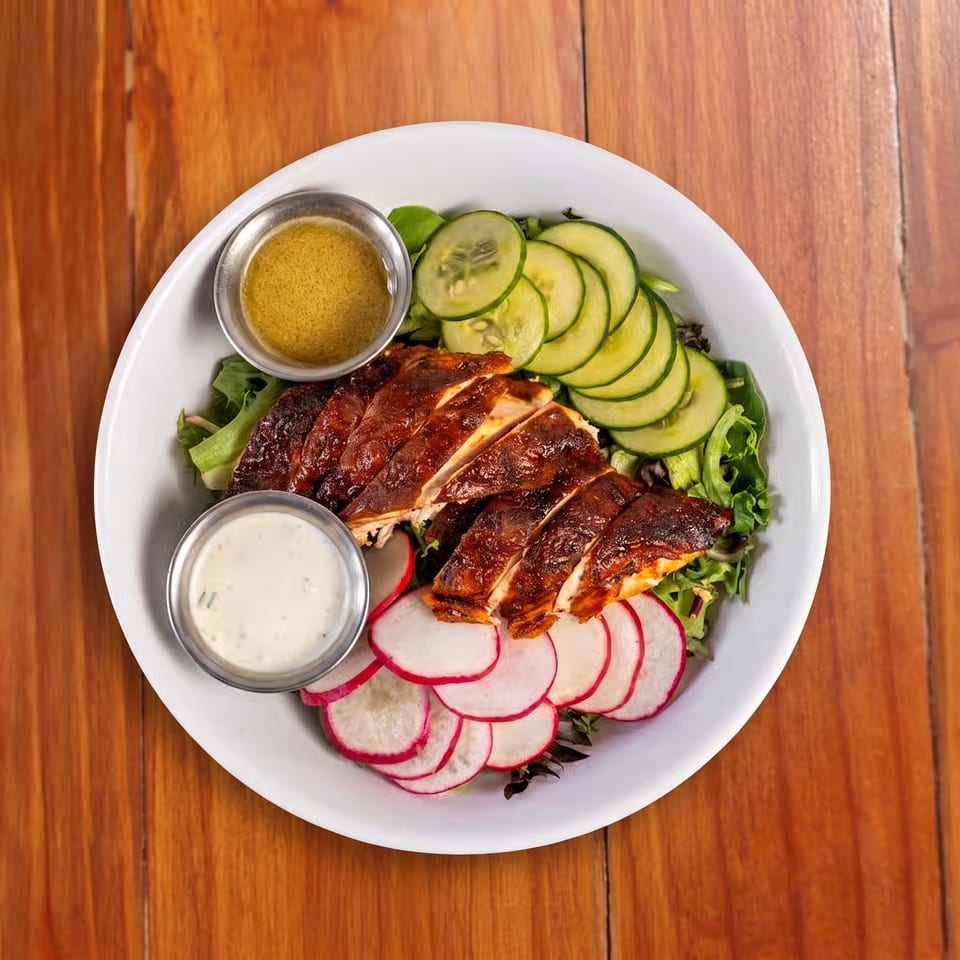 Sliced grilled chicken salad with cucumbers, radishes, greens, and two dipping sauces on a white plate.