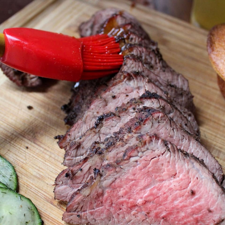Sliced medium-rare roast beef on a wooden cutting board with a red basting brush, bread, and cucumber slices.