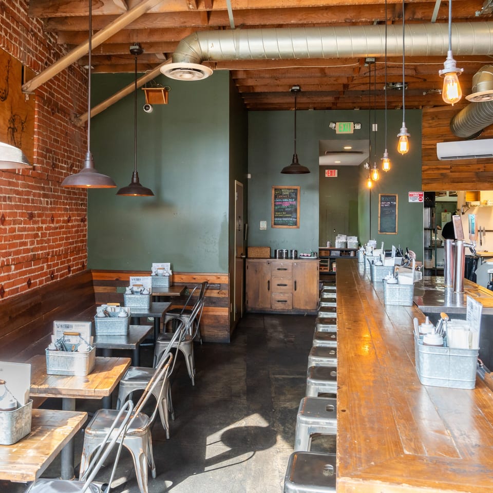 Rustic restaurant interior with exposed brick walls, wooden tables, metal stools, pendant lights, and a long bar counter.