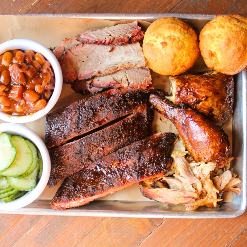 Barbecue platter with ribs, sliced brisket, pulled pork, chicken, cornbread, baked beans, and pickles on a metal tray.