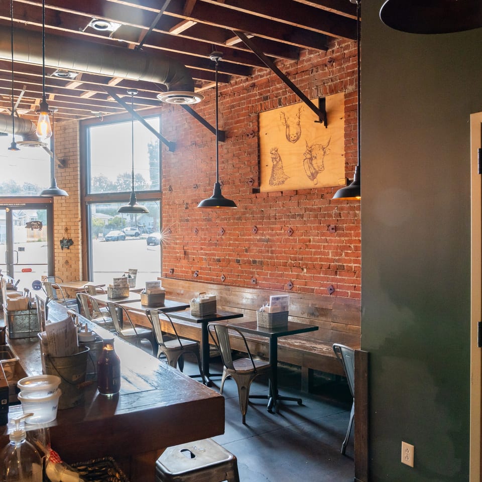 Rustic restaurant dining room with exposed brick walls, pendant lights, wooden tables, metal chairs, and large windows.