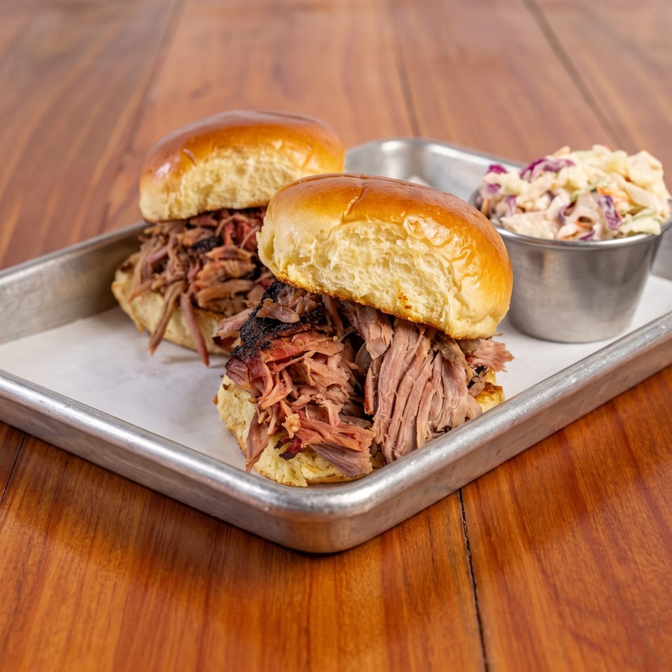 Pulled pork sandwiches on brioche buns with creamy coleslaw, served on a metal tray atop a wooden table.