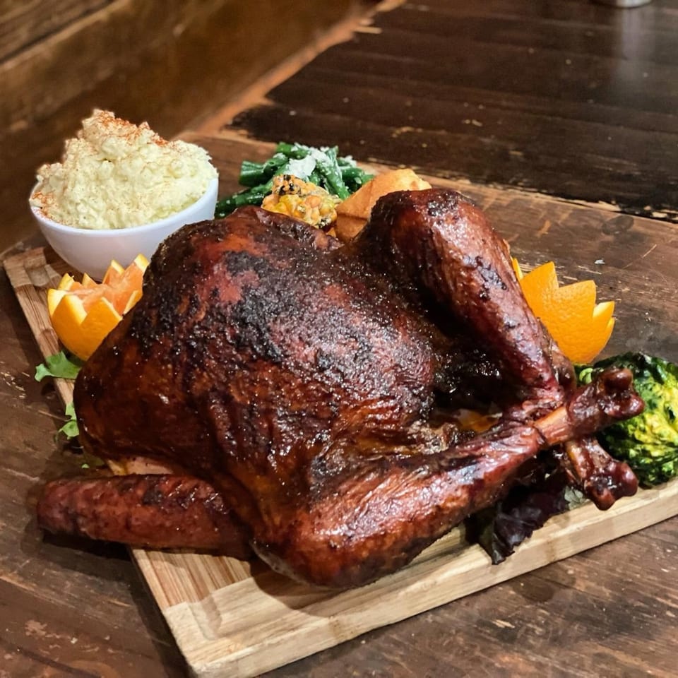 Glazed roasted turkey served on a wooden board with mashed potatoes, greens, vegetables, and orange wedges.