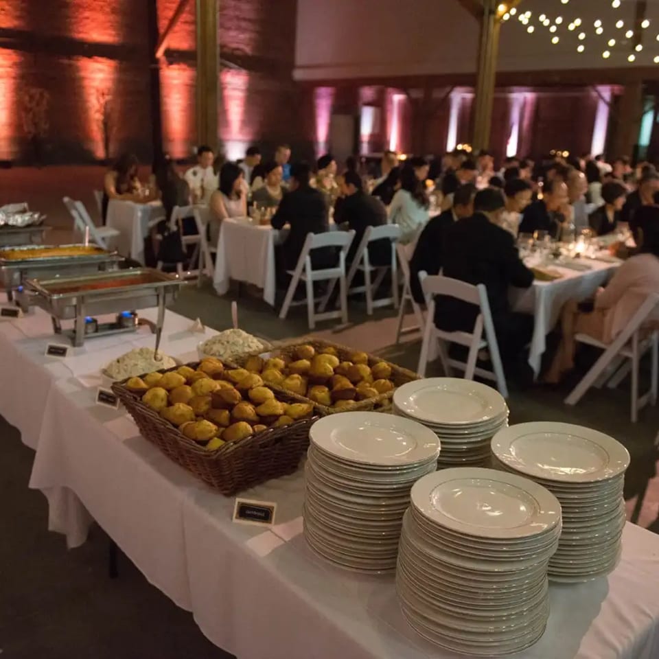 Buffet table with baskets of bread, chafing dishes, and stacked plates at a warmly lit event filled with seated guests.