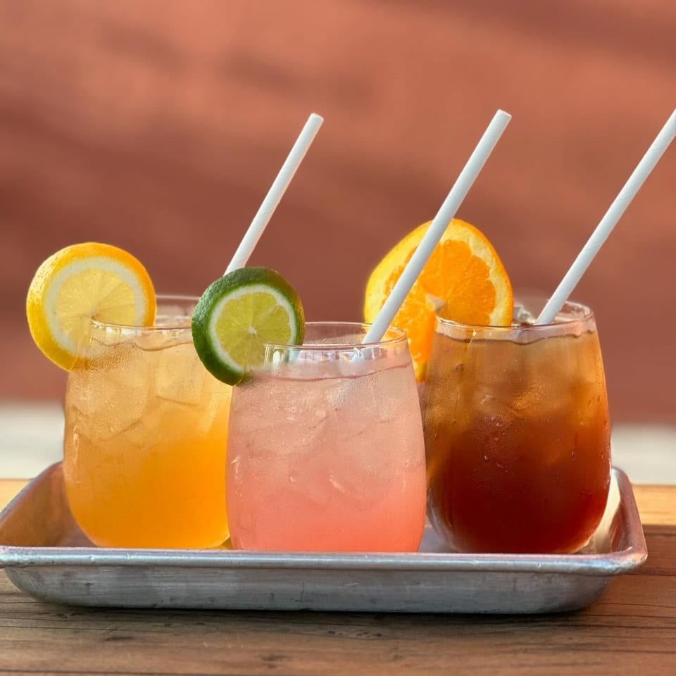 Three colorful iced drinks with citrus garnishes and white straws arranged on a metal tray.