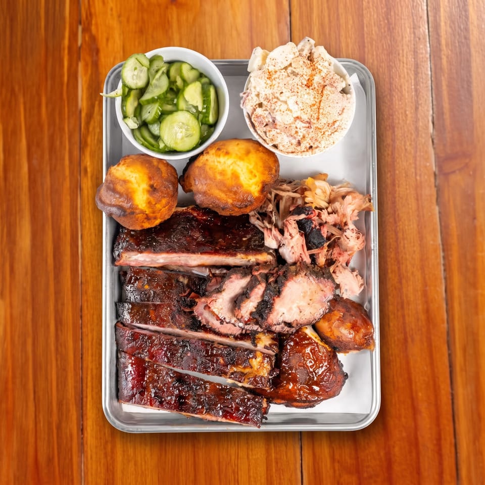 Overhead view of a barbecue platter with sauced ribs, pulled pork, cornbread, cucumber salad, and creamy coleslaw on a metal tray.
