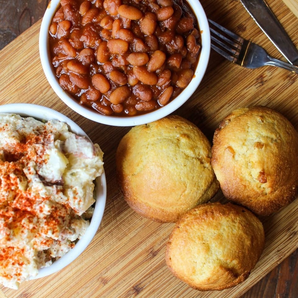 Overhead view of baked beans, paprika-dusted potato salad, and golden cornbread muffins arranged on a wooden serving board.