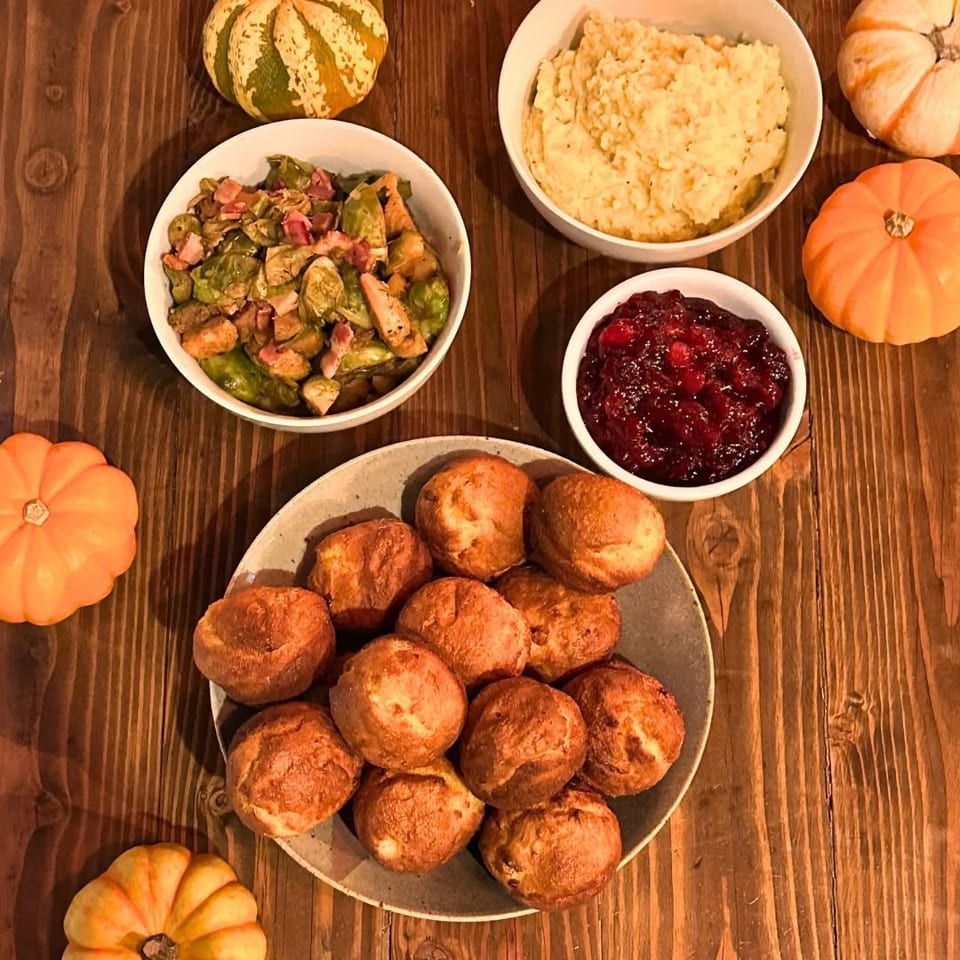 Overhead spread of mashed potatoes, Brussels sprouts with bacon, cranberry sauce, and dinner rolls surrounded by decorative pumpkins.
