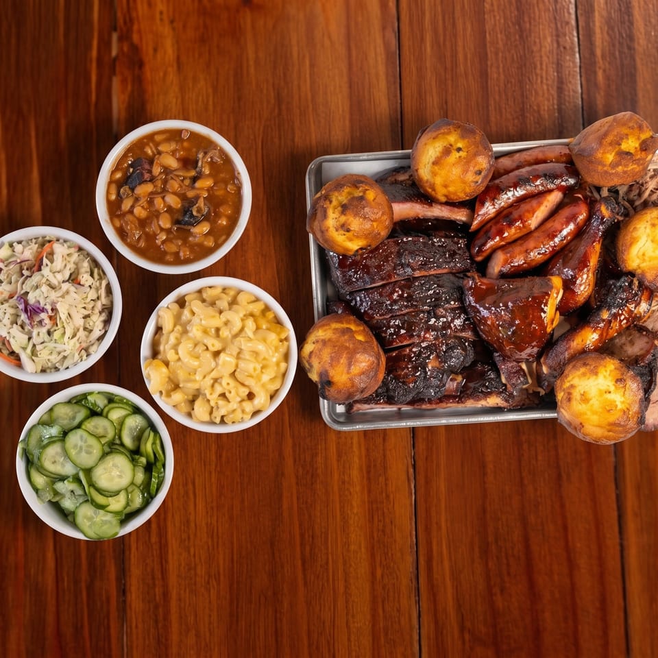 Overhead spread of barbecue ribs, sausages, cornbread, baked beans, coleslaw, macaroni and cheese, and cucumber salad on a wooden table.