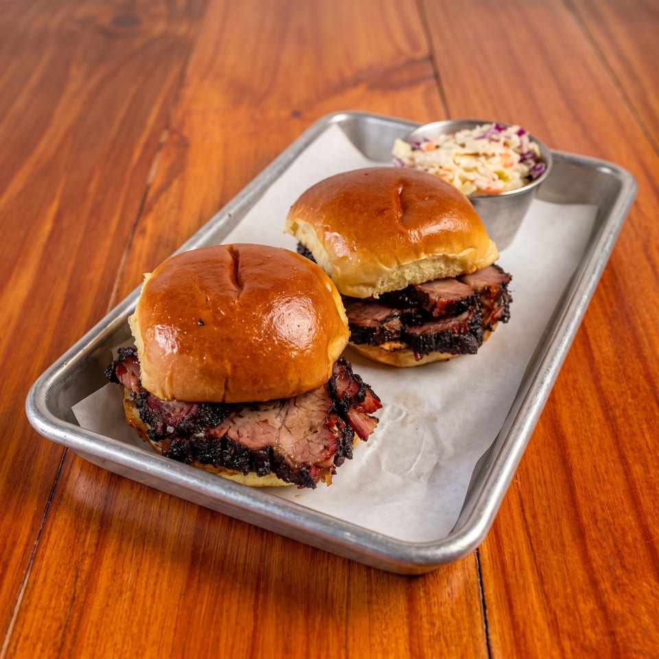 Two barbecue sandwiches on glossy buns sit in a metal tray with creamy coleslaw on a wooden restaurant table.