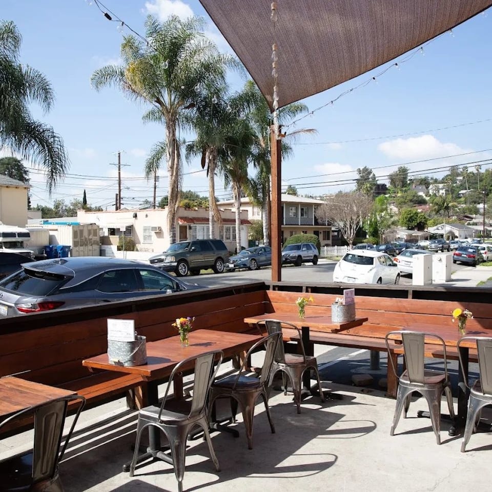 Sunny restaurant patio with wooden tables, metal chairs, flower vases, string lights, palm trees, and a neighborhood street view.