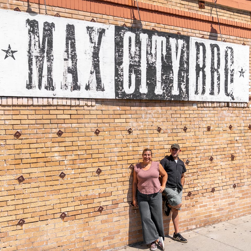 Two people pose beneath a weathered “Milk and Honey” sign on a sunlit brick restaurant wall.