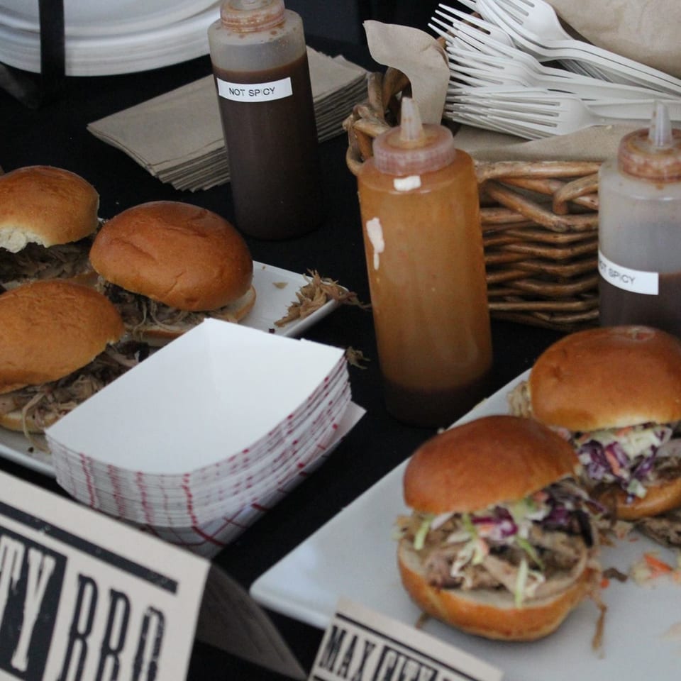 Pulled pork sliders with coleslaw arranged on trays beside labeled spicy and non-spicy sauce bottles, napkins, and plastic forks.