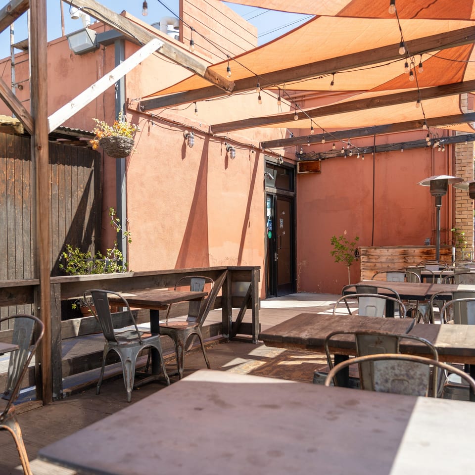 Sunlit restaurant patio with wooden tables, metal chairs, orange shade sails, string lights, and outdoor heaters.