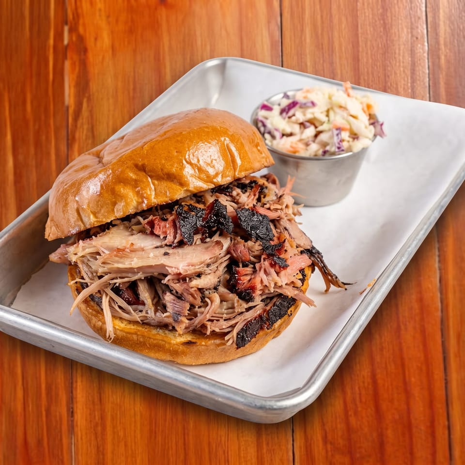 Pulled pork sandwich on a glossy bun with charred meat, served alongside creamy coleslaw on a metal tray.