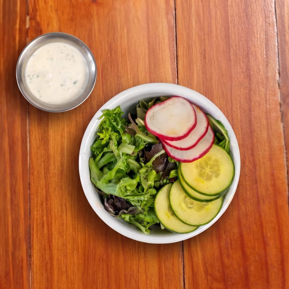 Bowl of mixed green salad topped with sliced cucumbers and radishes, accompanied by creamy dressing on a wooden table.