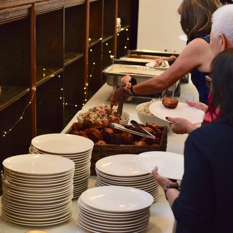 Guests serve themselves from a buffet featuring roasted meat, side dishes, and stacks of white plates in a warmly decorated dining area.